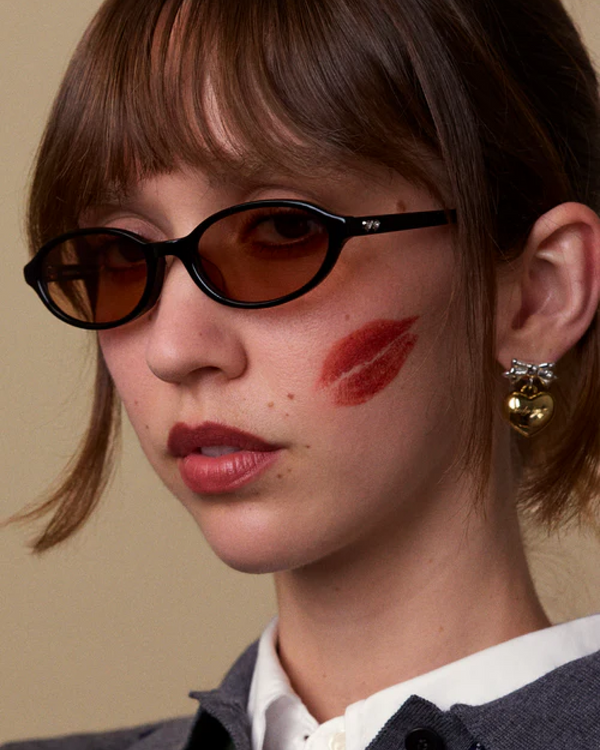 Model wearing Bonnie Clyde Baby sunglasses with black oval frames and brown-tinted lenses, close-up portrait